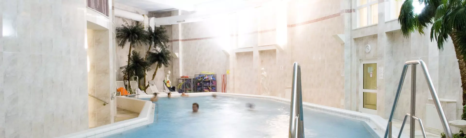Innenansicht eines hellen Therapiebads mit warmem Licht, Palmen-Dekoration, flacher Einstiegstreppe und mehreren Personen im Wasser.